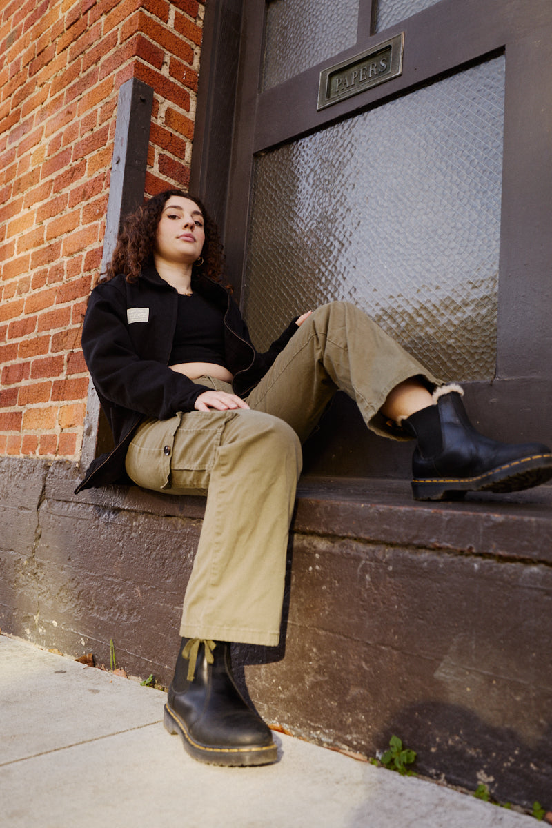 A person with curly hair sits on a stoop by a brick wall, wearing rugged workwear: boots, khaki pants, and the classic Bridge Co. Chore Jacket made in San Francisco.