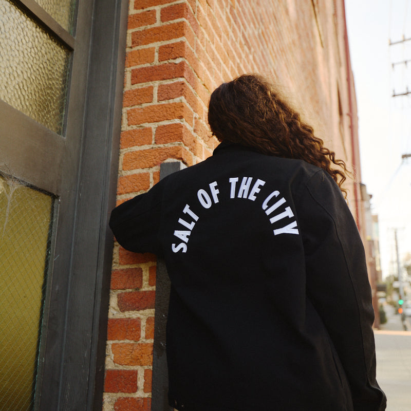 A person with curly hair stands by a brick wall wearing the Bridge Co. Chore Jacket in black, featuring "SALT OF THE CITY" text and made in San Francisco.