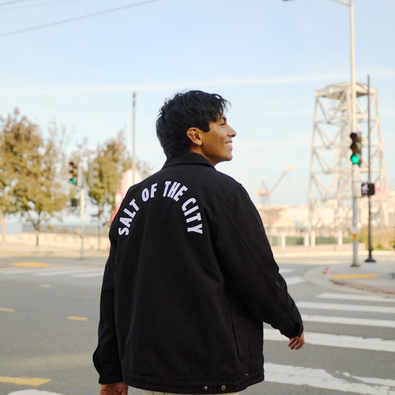 A person crosses the street in a black Bridge Co. Chore Jacket with SALT OF THE CITY on the back, capturing classic workwear style made in San Francisco.