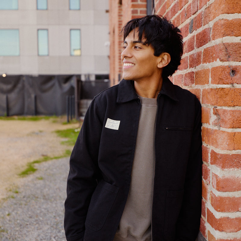 A young man in a Bridge Co. Chore Jacket smiles as he leans against a red brick wall outdoors, highlighting classic workwear style.