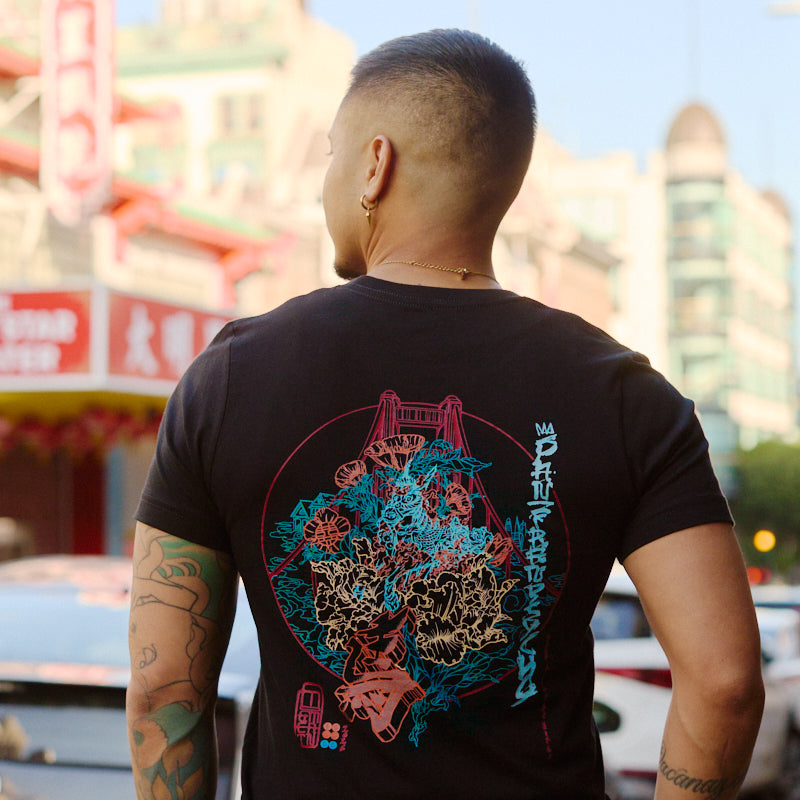 A tattooed man stands on a city street with colorful buildings, wearing the Year of the Horse Tee—a black graphic t-shirt made from soft Airlume cotton.