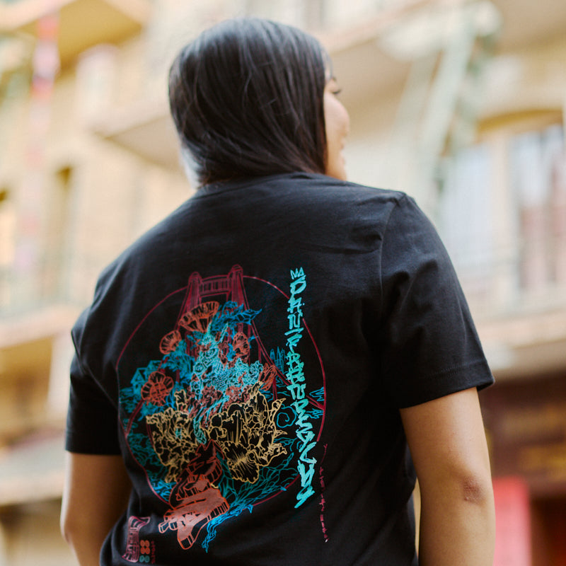 Wearing the Year of the Horse Tee—a black Airlume cotton T-shirt with colorful abstract graphics—standing outdoors near buildings, perfect for celebrating Lunar New Year.