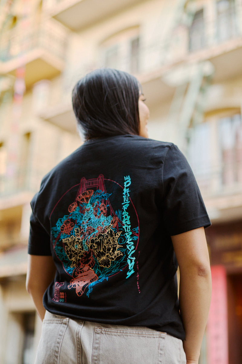 Wearing the Year of the Horse Tee—a black Airlume cotton T-shirt with colorful abstract graphics—standing outdoors near buildings, perfect for celebrating Lunar New Year.