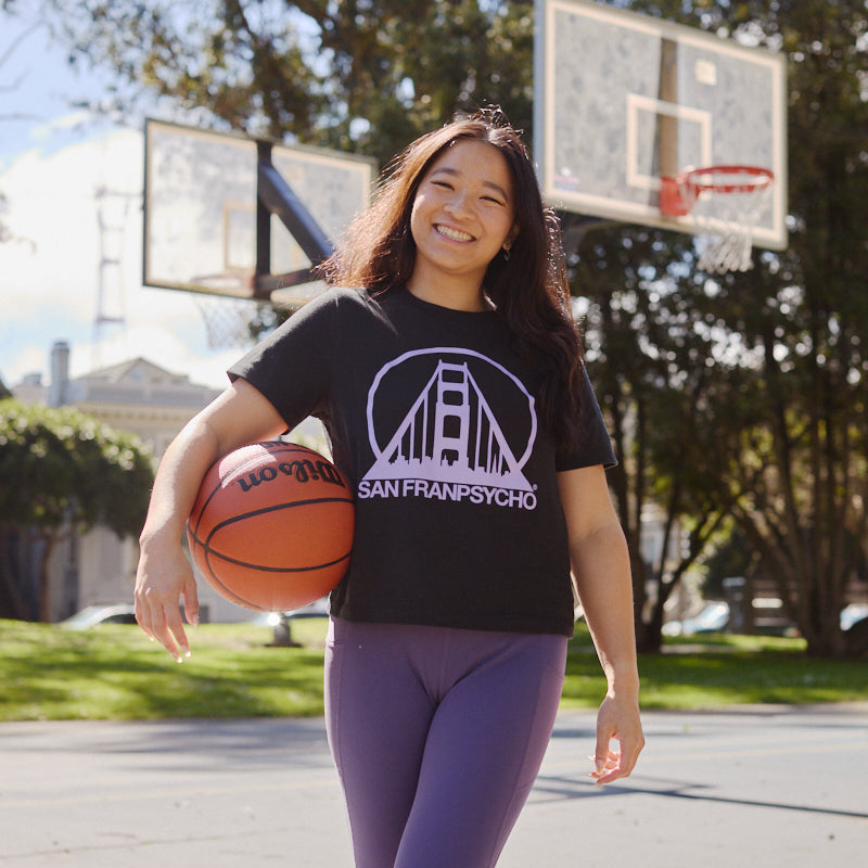 Smiling woman holding a basketball on an outdoor court, wearing the Women's Black & Purple Logo Crop Tee and purple leggings.