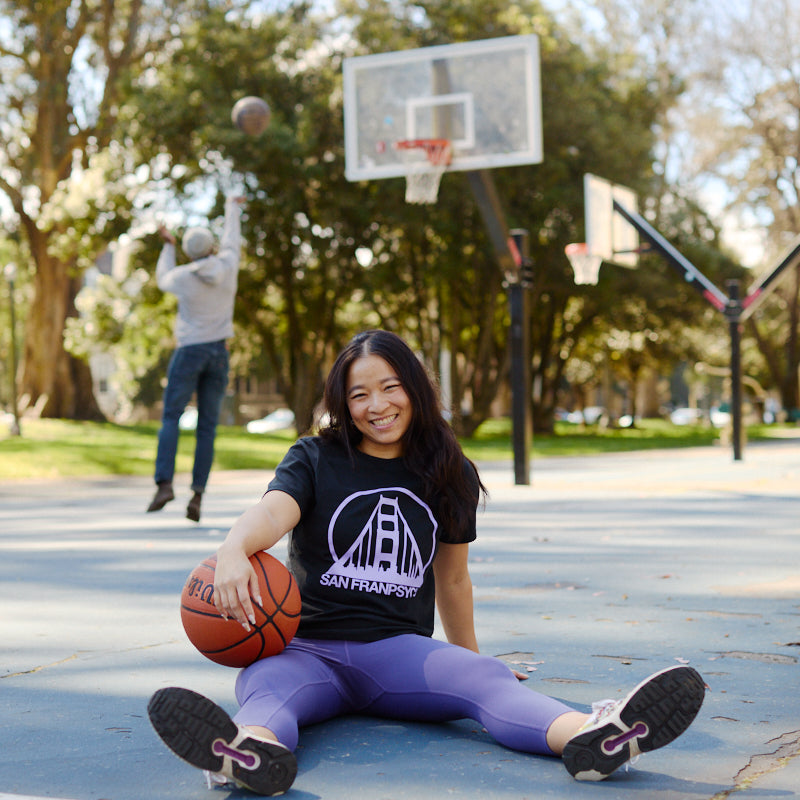 A young woman in a Women's Black & Purple Logo Crop Tee sits on an outdoor basketball court, smiling with a basketball, as someone shoots a basket behind her.