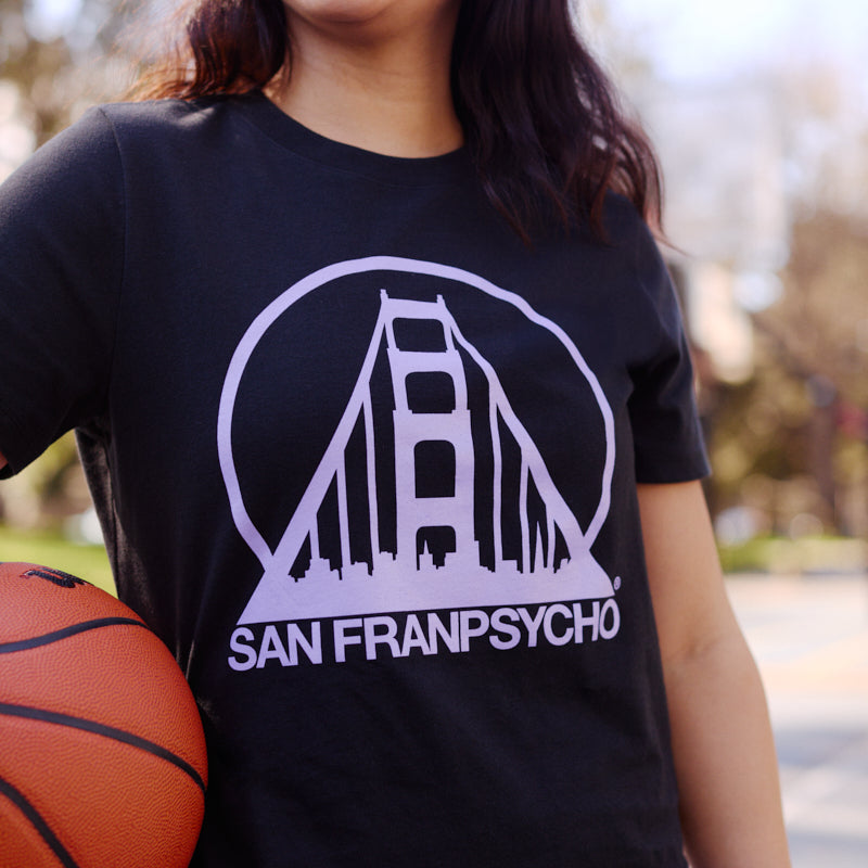 Smiling woman outdoors holding a basketball, wearing a Women's Black & Purple Logo Crop Tee.