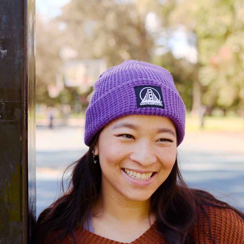 A woman wearing the Purple Waffle Beanie w/ Black/White Logo and a brown sweater leans against a pole outdoors on a sunny day.