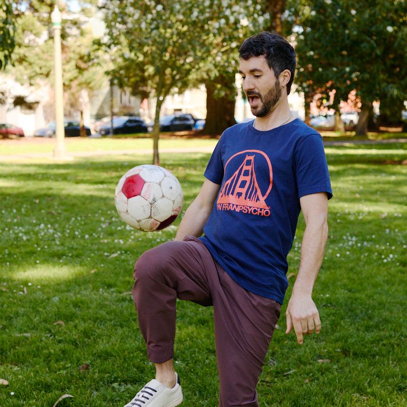 A man in a Navy & Poppy Logo Tee balances a soccer ball on his knee in a sunny park with trees in the background.