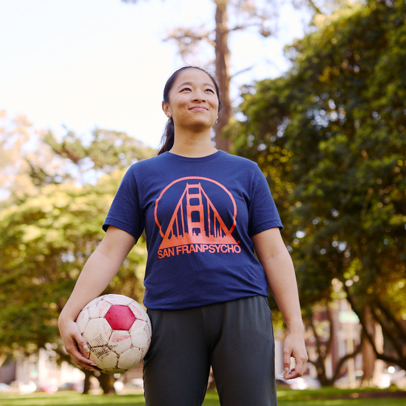 Person wearing a Navy & Poppy Logo Tee holds a soccer ball while standing on grass in a sunny park.