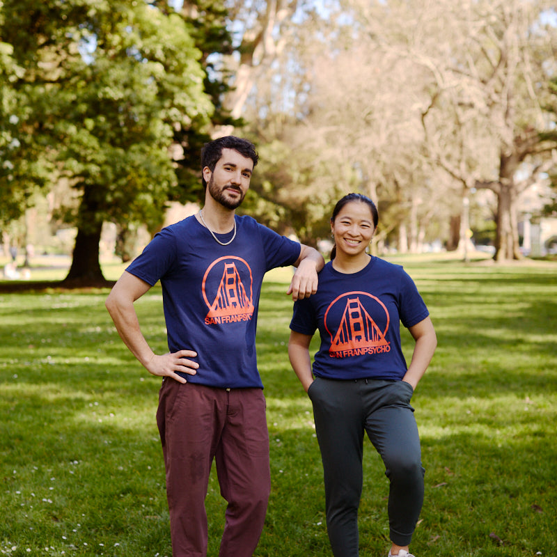 Two people wearing matching Navy & Poppy Logo Tees pose on grass in a park, one resting a hand on a soccer ball.