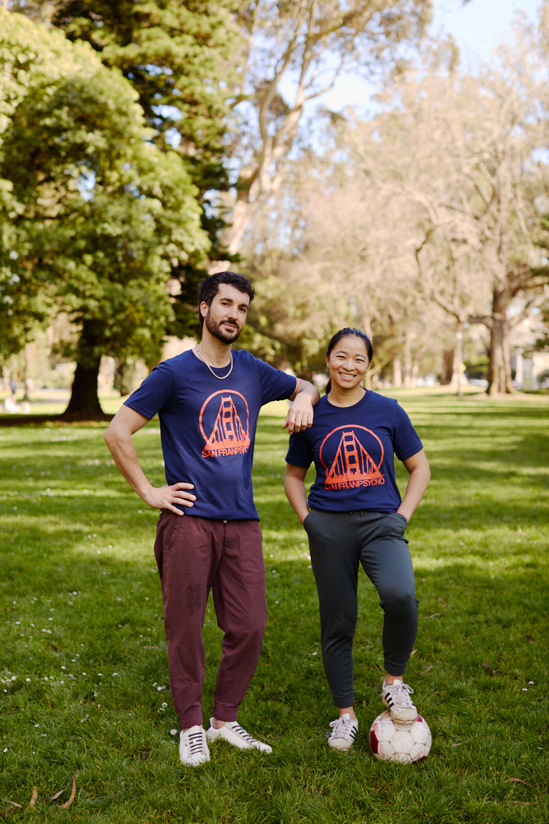 Two people wearing matching Navy & Poppy Logo Tees pose on grass in a park, one resting a hand on a soccer ball.