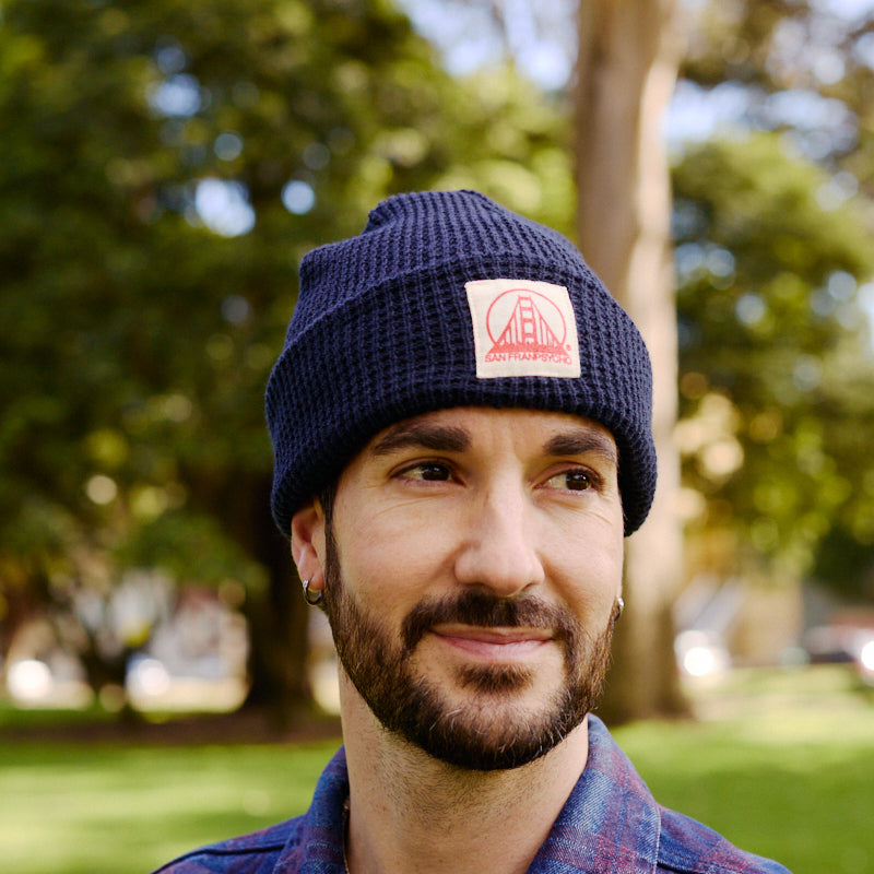 A bearded man in a plaid shirt stands outside in a sunny, green park, wearing a Navy Waffle Beanie with Natural/Poppy Logo.