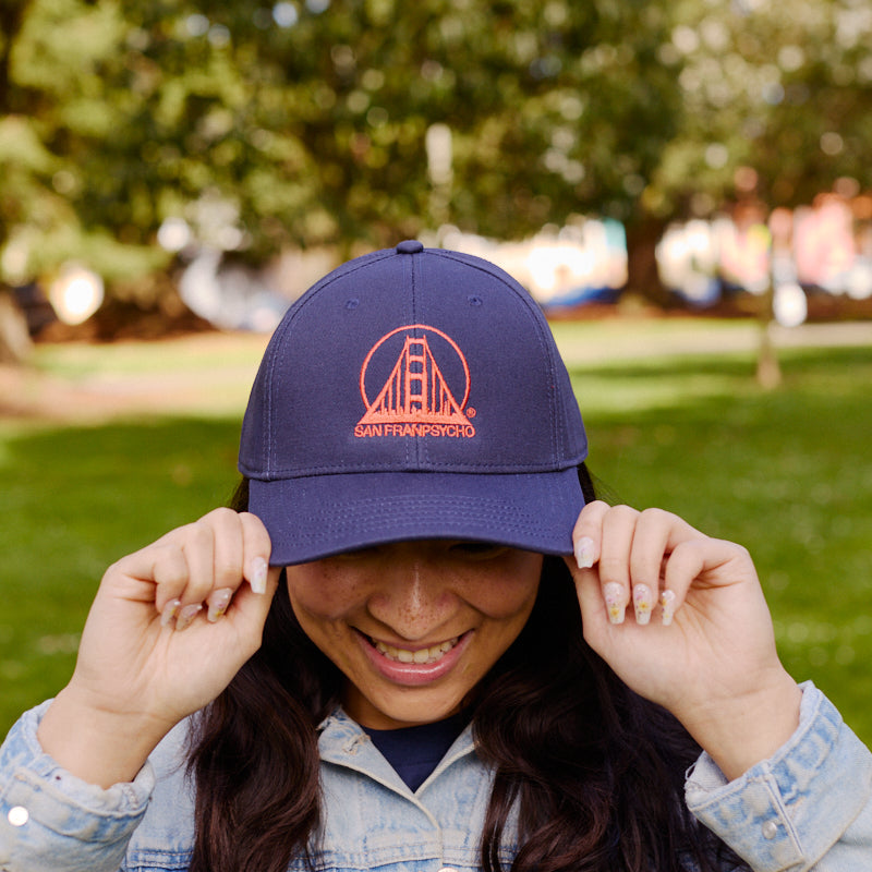 A smiling person in a denim jacket holds the Embroidered Logo Snapback - Navy & Poppy outdoors in a sunny, green park.