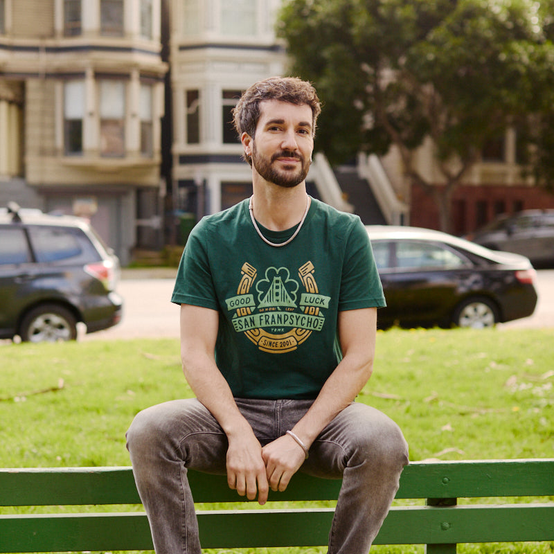 A man in a Lucky Psycho Tee sits on a green bench in a park, with houses and trees in the background.