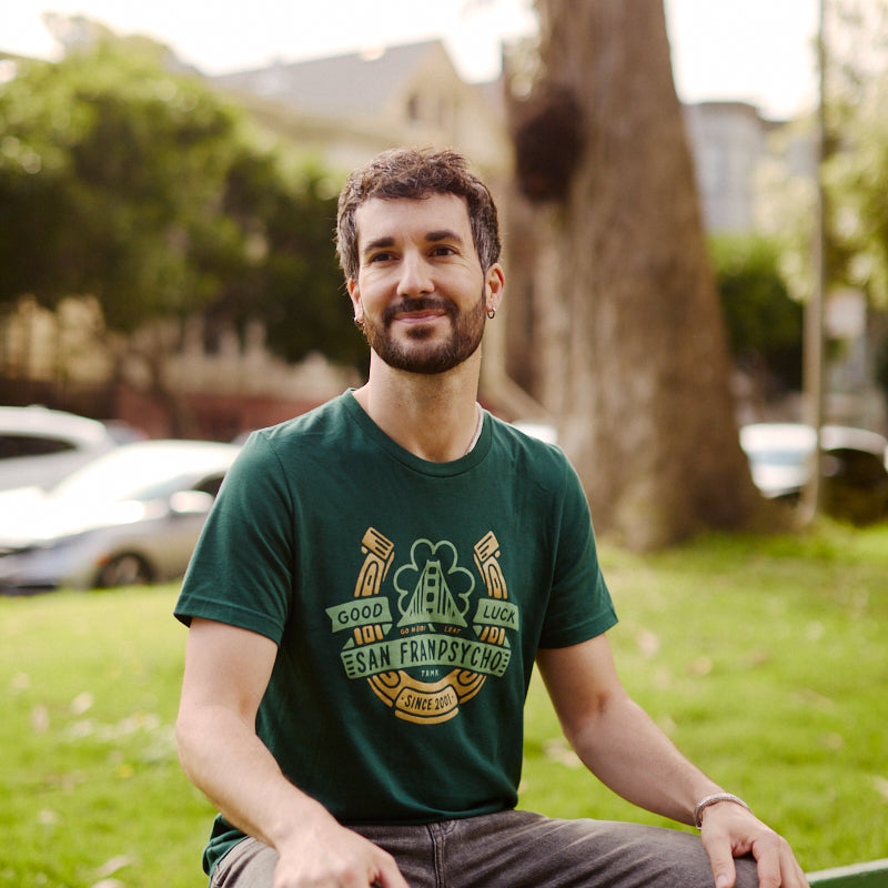 Man with short dark hair and a beard sits on a green park bench, wearing the Lucky Psycho Tee, a hand-printed St. Paddy’s shirt featuring San Francisco.