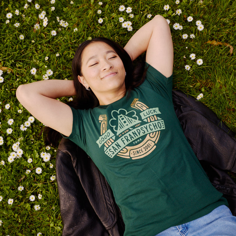 Woman lies on grass among white flowers, smiling with eyes closed in a hand-printed Lucky Psycho Tee and blue jeans—celebrating St. Patrick’s Day in style.