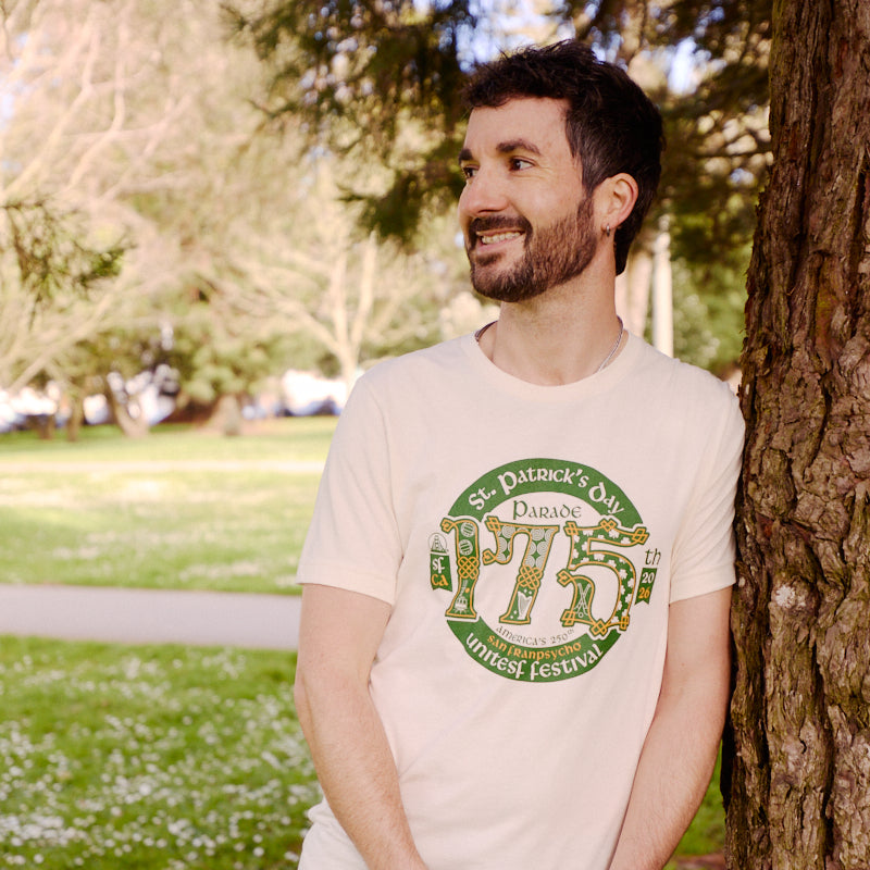 Man smiling and leaning against a tree in a park, wearing the 175th St Patrick's Day Parade Tee, a white limited edition commemorative shirt designed for the festival.