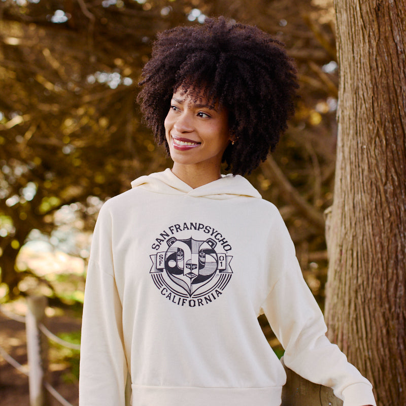 A woman in a Women's Bear Essentials Pullover and jeans smiles outdoors by a tree, sunlight streaming behind her.