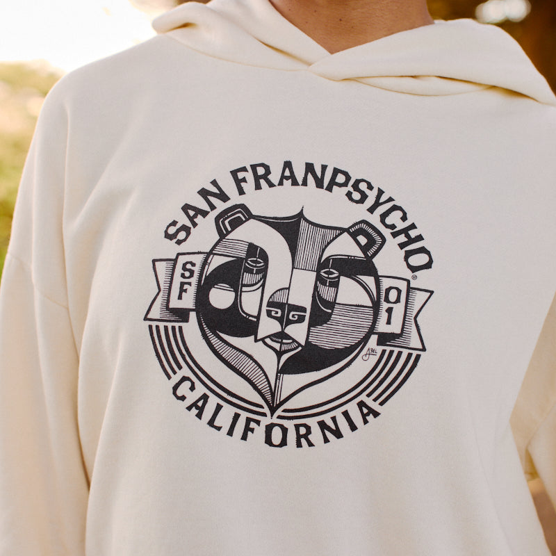 A smiling woman wears the Women's Bear Essentials Pullover, an ultra-soft cream hoodie featuring “San Franpsycho California” and a panda logo.