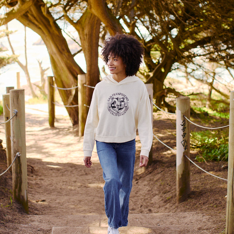 A person with curly hair walks along a sandy path lined with trees, wearing jeans and the ultra-soft Women's Bear Essentials Pullover.