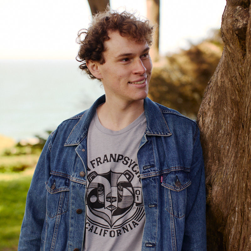 A young man in a denim jacket leans against a tree, smiling, with his Bear Essentials Tee in Heather Grey peeking out—perfect for relaxing by the greenery and water.