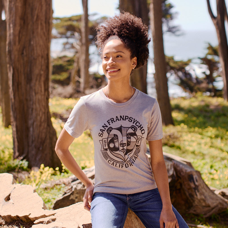A woman smiles outdoors in a forest, wearing the ultra-soft Bear Essentials Tee - Heather Grey with a California bear graphic and jeans.