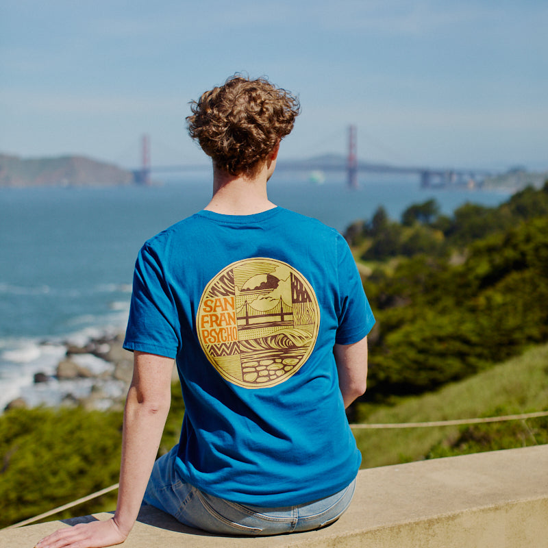 Wearing the Elemental Tee - Blue, a person sits on a ledge overlooking the Golden Gate Bridge and San Francisco Bay on a sunny day.