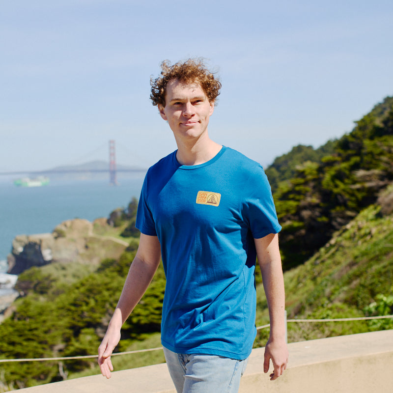 A young man wearing the Elemental Tee - Blue strolls outdoors with the Golden Gate Bridge and lush greenery in the background, embodying San Francisco’s relaxed vibe.