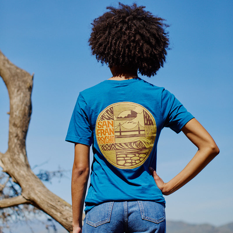 Person wearing the Elemental Tee - Blue, featuring a San Francisco graphic, stands outdoors facing away near a tree and blue sky. Crafted from 100% cotton for comfort.
