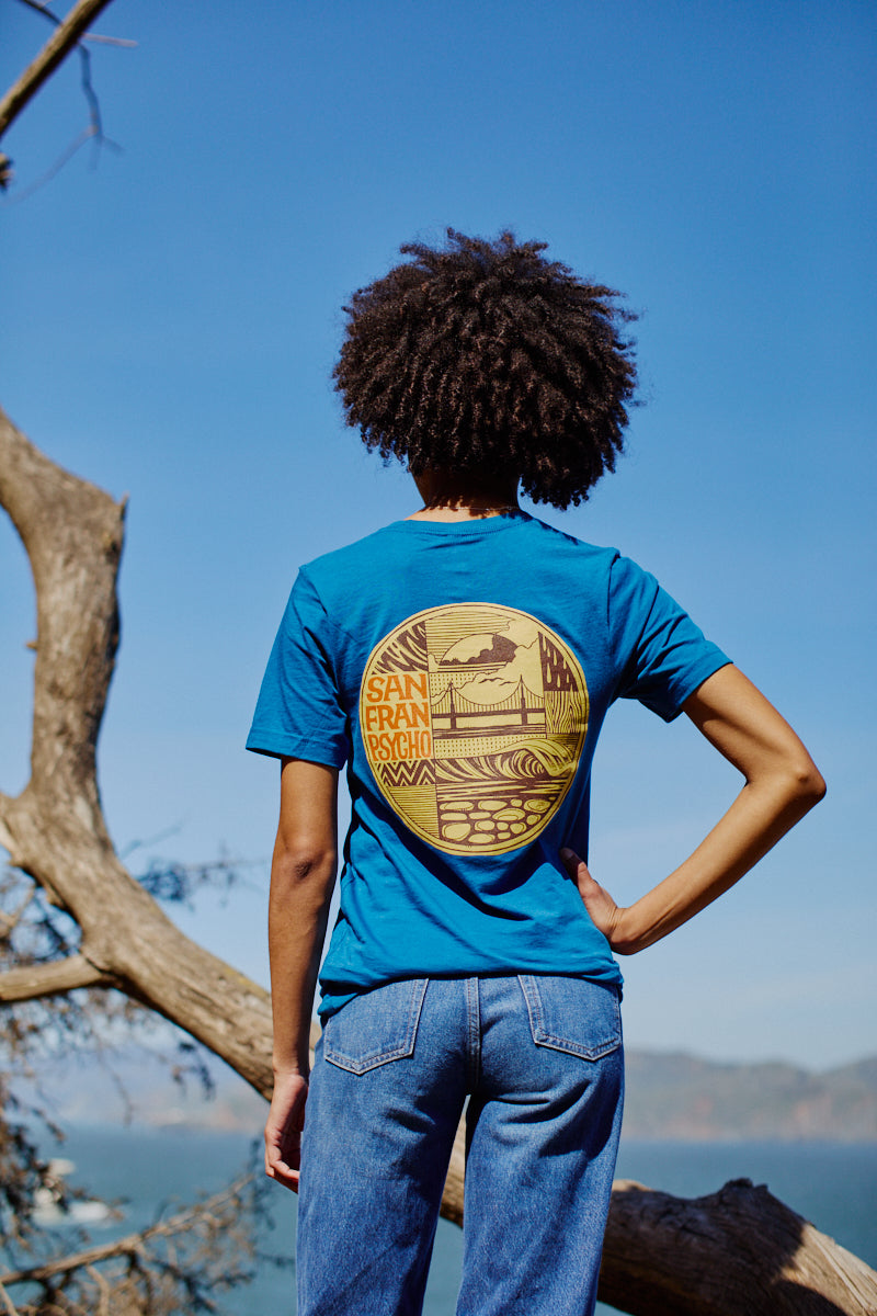Person wearing the Elemental Tee - Blue, featuring a San Francisco graphic, stands outdoors facing away near a tree and blue sky. Crafted from 100% cotton for comfort.