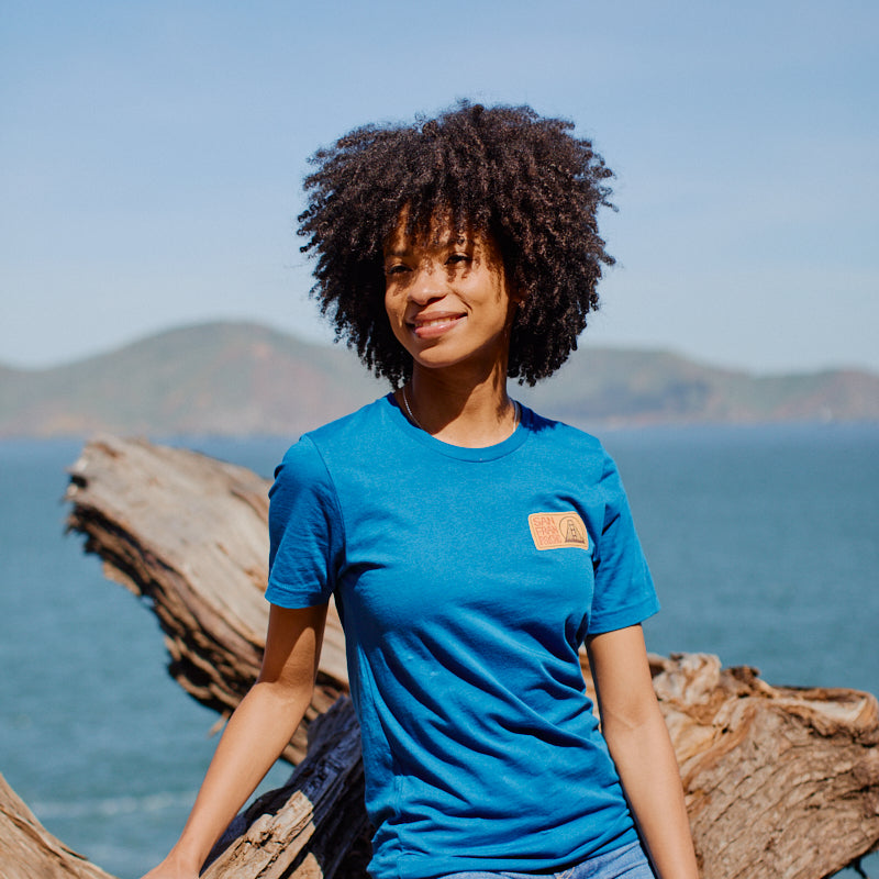 A woman in the Elemental Tee - Blue and jeans sits on driftwood by the sea, with San Francisco hills and a clear sky in the background.