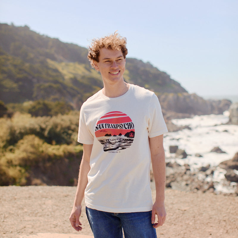 A smiling person in jeans and a Kelly's Cove Tee - Vintage White stands outdoors by the ocean and cliffs, enjoying the sunny coastal city vibes.