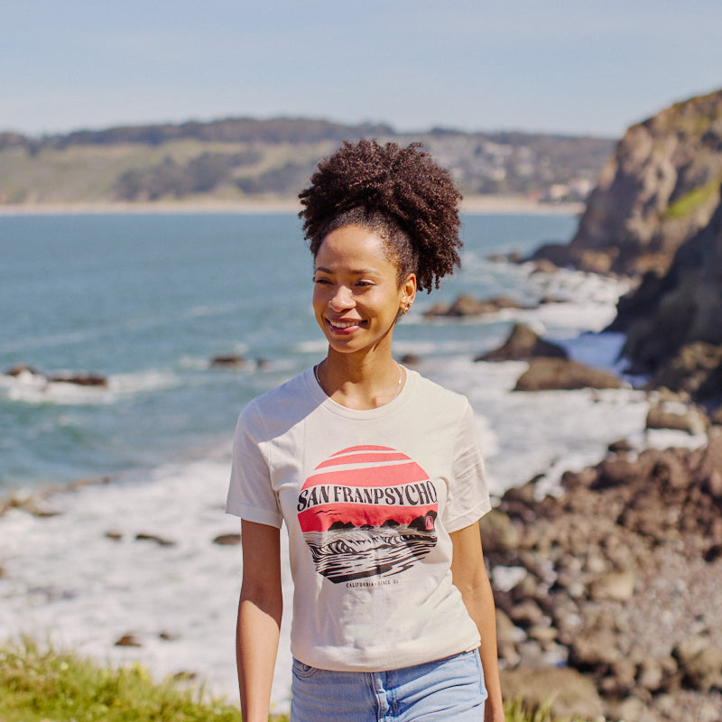 A woman in the Kelly's Cove Tee - Vintage White strolls along a rocky coastline, ocean and cliffs behind her—capturing relaxed coastal city style.