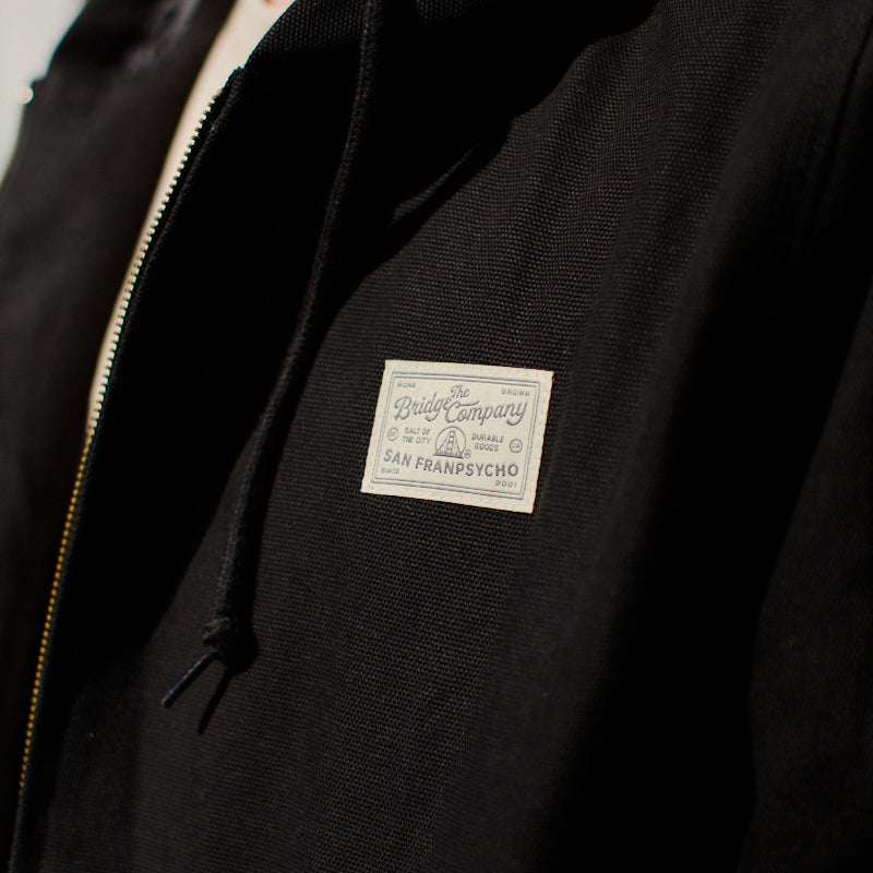 Close-up of the Bridge Co. Workwear Jacket in black cotton canvas, featuring a zip-up front and a white patch that reads The Daily Company San Francyscho.
