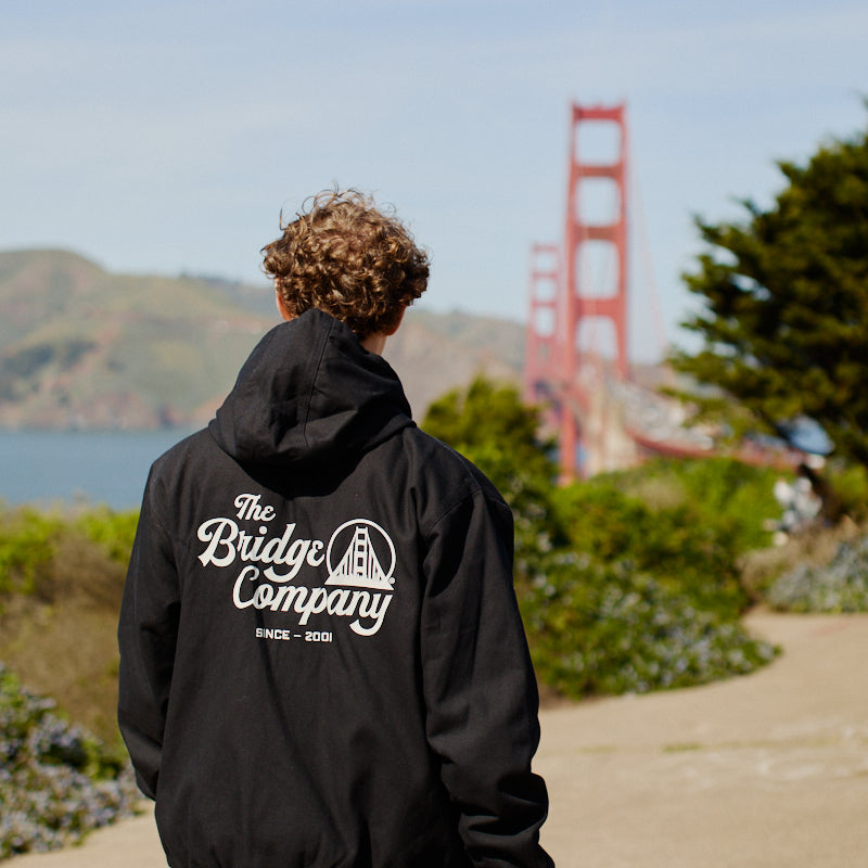 A person in a black hoodie and the Bridge Co. Workwear Jacket walks near the Golden Gate Bridge in daylight.