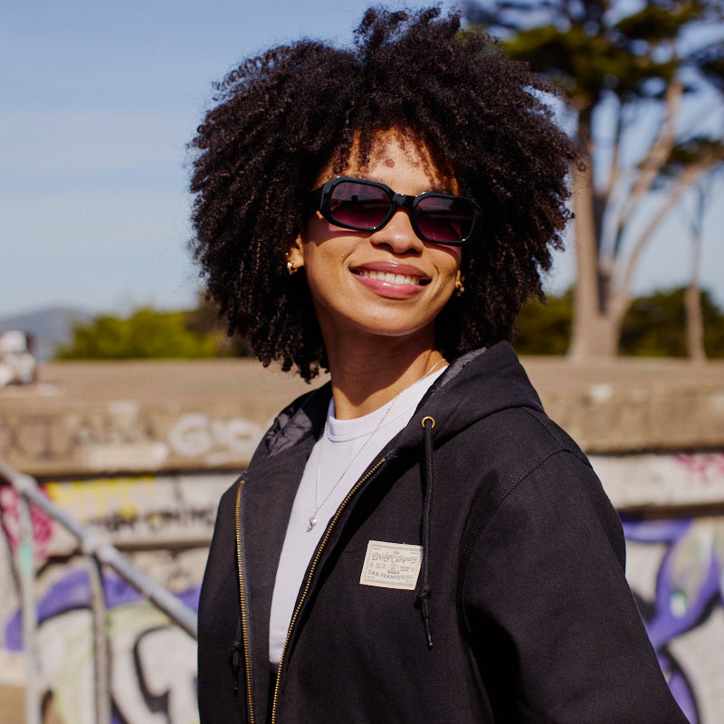 A smiling woman with curly hair and sunglasses stands outdoors by a graffiti wall and trees, wearing the Bridge Co. Workwear Jacket.