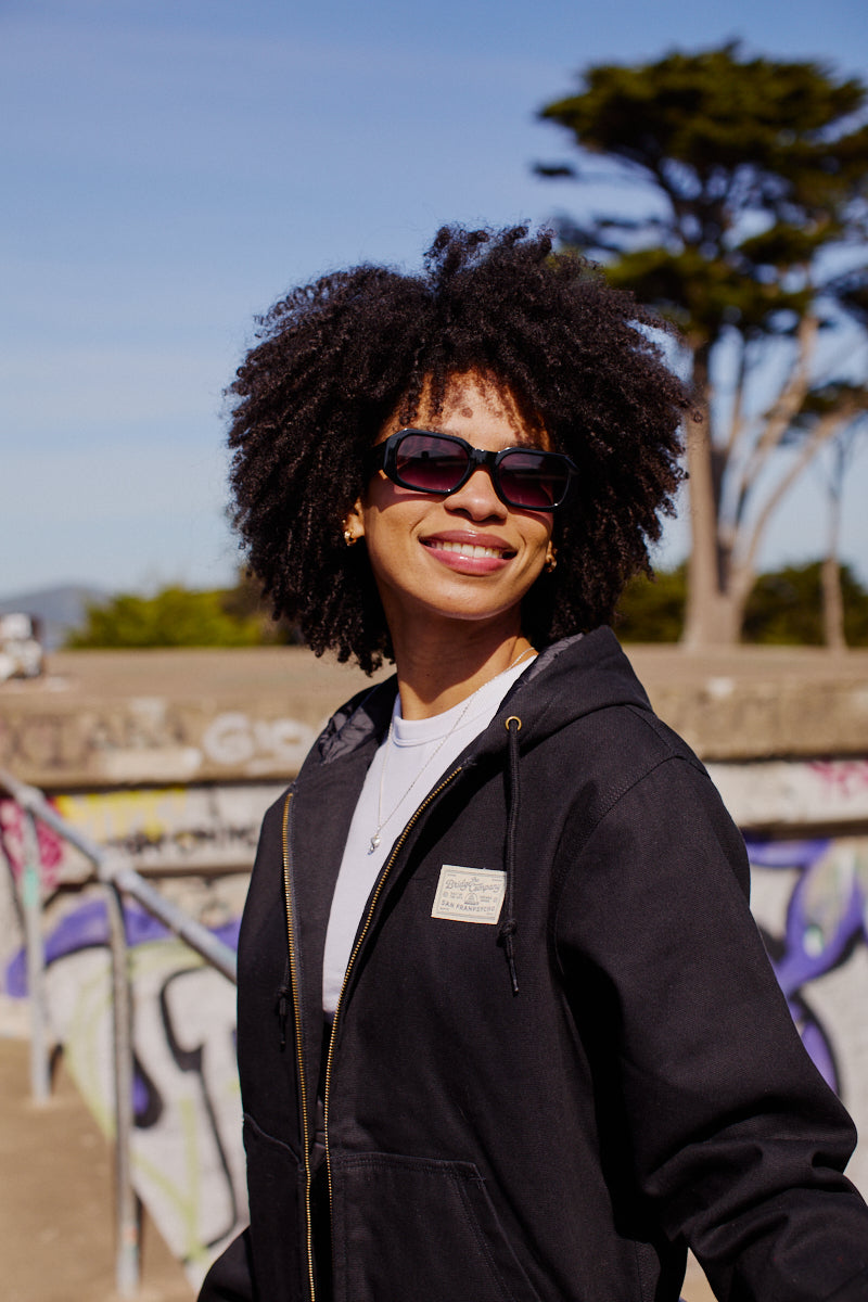 A smiling woman with curly hair and sunglasses stands outdoors by a graffiti wall and trees, wearing the Bridge Co. Workwear Jacket.