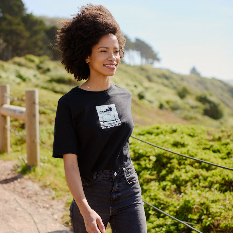 A smiling woman strolls along a scenic, sunlit path in greenery and distant trees, wearing the Women's Elemental Cropped Tee in black with its bold graphic design.