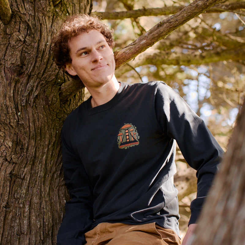 A person with curly hair sits on a tree branch, wearing the Mind Bloom Long Sleeve inspired by Erik Abel artwork and tan pants.