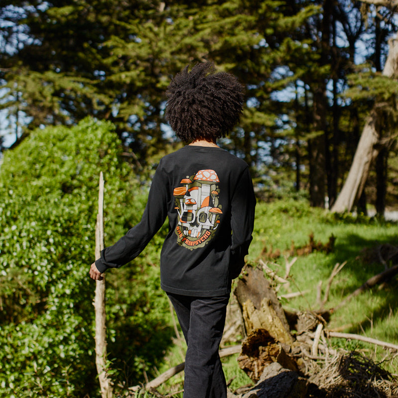 Wearing the Mind Bloom Long Sleeve, a person in black walks on logs through a sunlit, green forest with tall trees in the background.