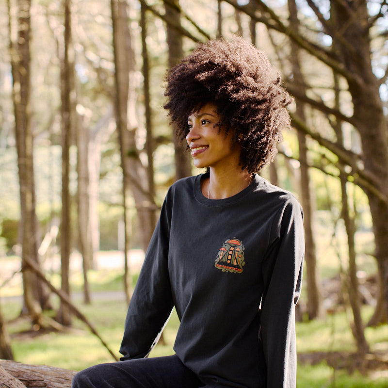 A woman with curly hair smiles while sitting on a fallen tree in the forest, wearing the Mind Bloom Long Sleeve, a graphic tee inspired by Erik Abel's artwork.