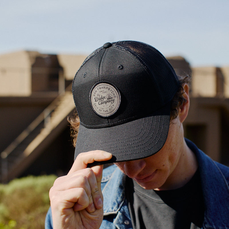 Outdoors on a sunny day, a person in a denim jacket tips the Salt of The City Trucker Hat—a black hat featuring a round patch and adjustable back.