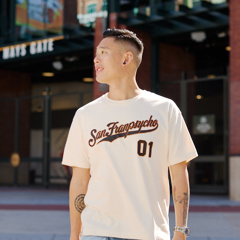 A man in a Psychos Script Tee - Natural stands outside a stadium gate, looking to the side.