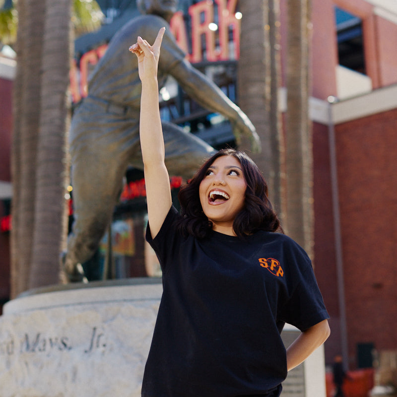 A woman smiles and points up in front of a giant baseball player statue outside the stadium, proudly wearing the Black & Orange Psychos Tee.