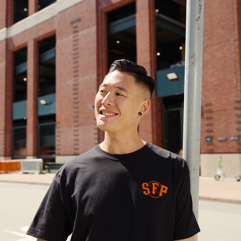 A person smiles while standing outside near a brick building and a street sign, wearing the Black & Orange Psychos Tee.