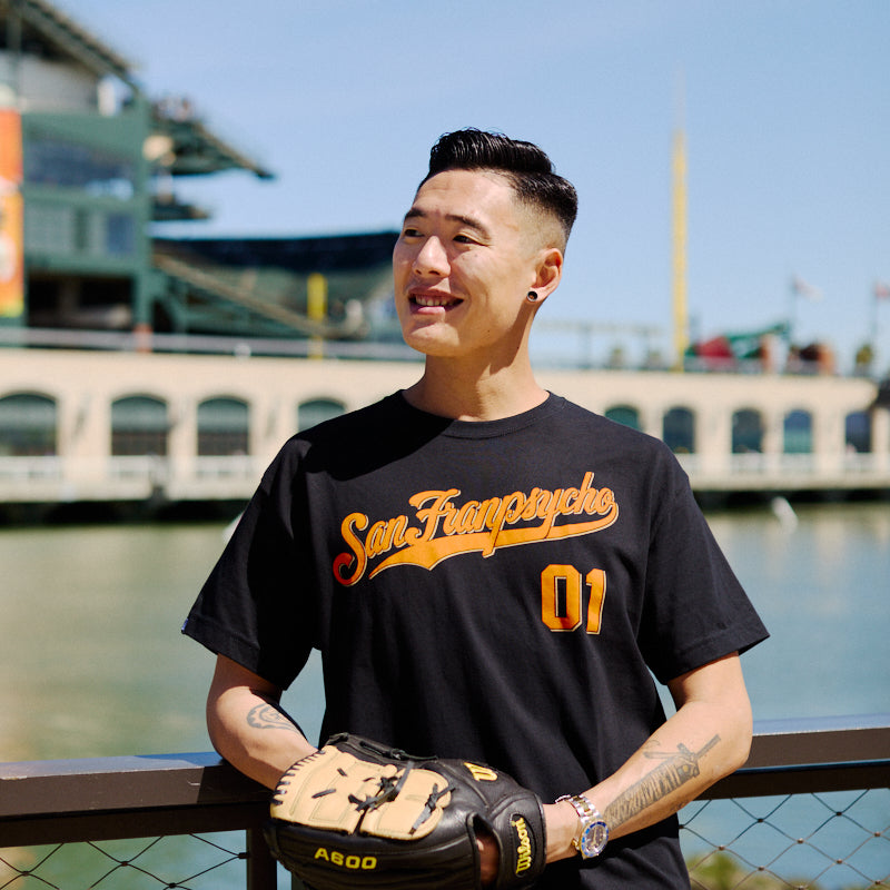 A man wearing the Psychos Script Tee - Black, featuring hand-printed orange graphics, holds a baseball glove while standing by a waterfront stadium.