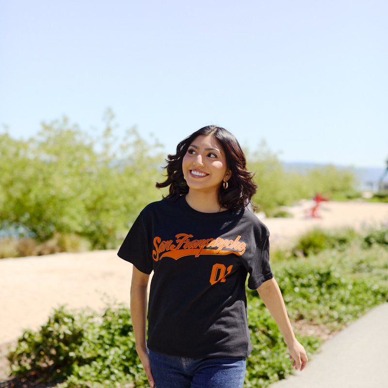 A woman smiles while walking outdoors on a sunny day, wearing the Psychos Script Tee - Black with a hand-printed San Francisco 01 design in black and orange, paired stylishly with jeans.