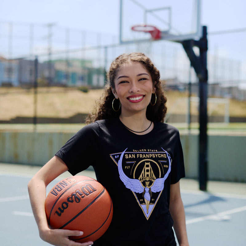 A smiling young woman wears a Valhalla Tee while holding a basketball on an outdoor court, with a hoop and net in the background—show your Valkyrie spirit this season with the Valhalla Tee.