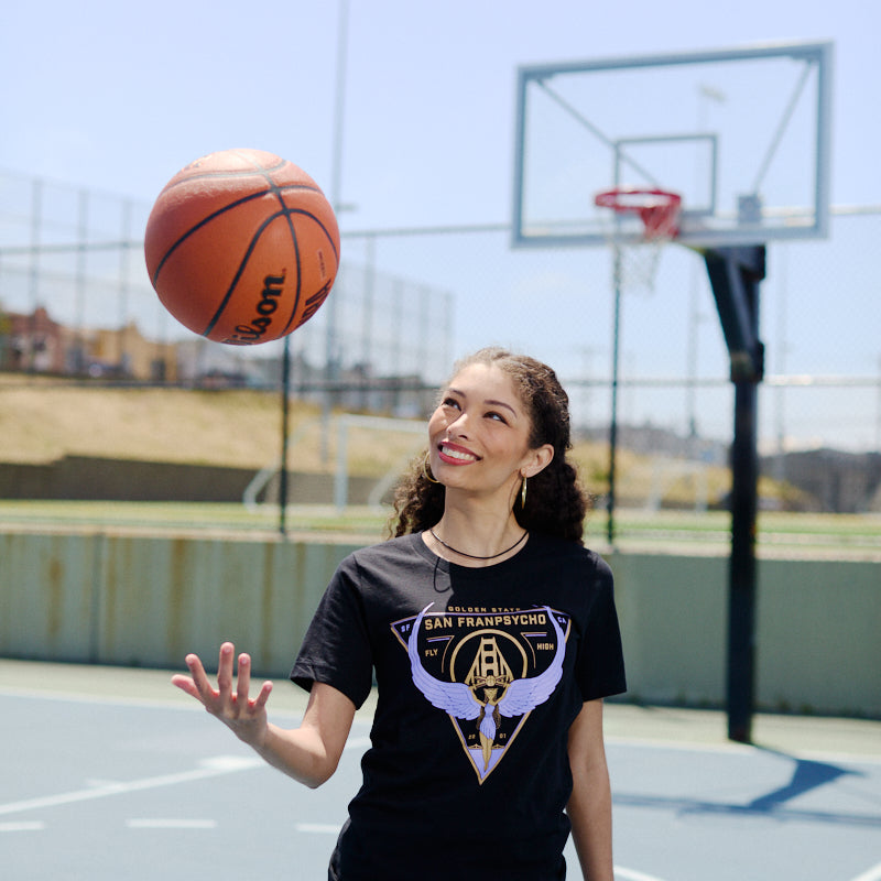 A woman smiles and spins a basketball on her finger at an outdoor court, wearing the Valhalla Tee—crafted from soft Airlume cotton and ideal for showing your Valkyries spirit this season.