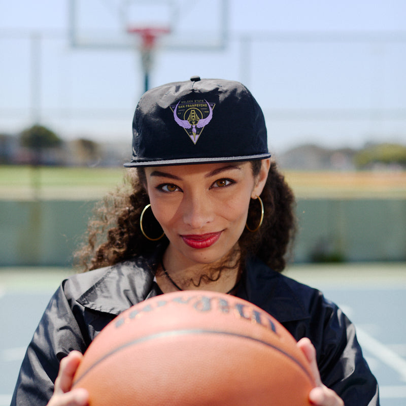 Woman in black Valhalla Hat with adjustable back and jacket holding a basketball on an outdoor court, basketball hoop visible in the background.